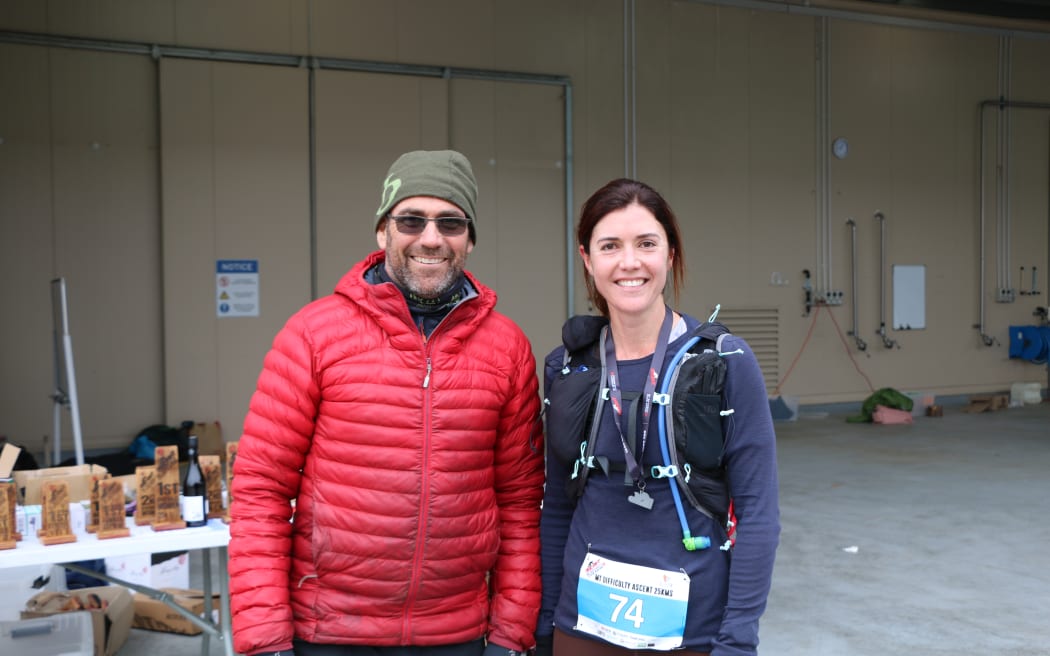 A man and a woman stand in front of a work building, smiling. The man is wearing a bright red puffer jacket and a khaki beanie. The woman is wearing a race number affixed to her blue long-sleeved shirt and has a backpack and Camelbak on.