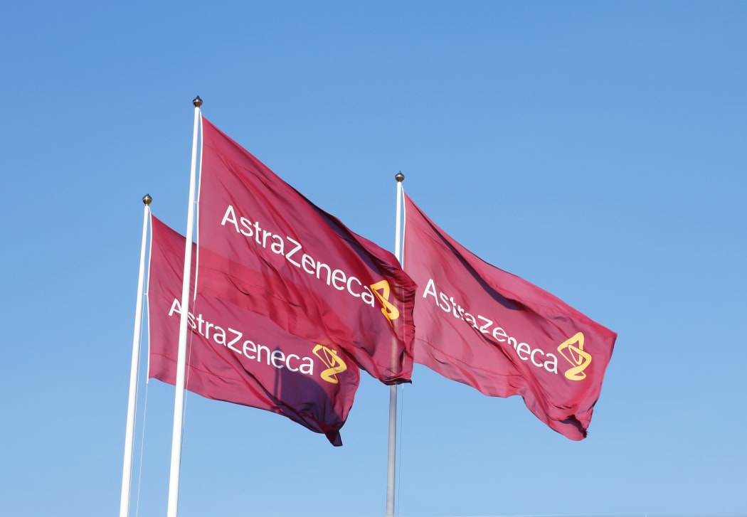 Three purple flags with the logo for Atrazeneca flying in the wind on top of the flagpoles against the blue sky.