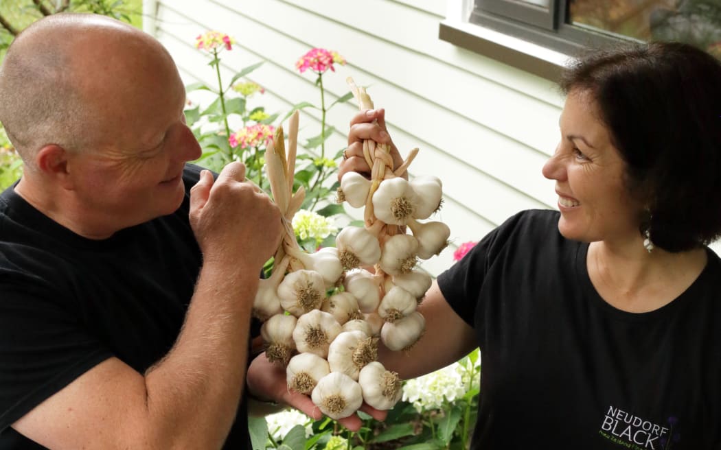 Noël and Teena sit on a bench outside holding a braid of garlic between them and smiling.