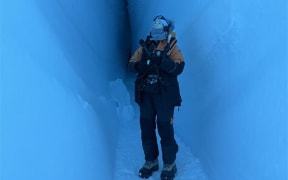 Holding microphones and kitted out in Antarctica New Zealand cold weather gear Claire is standing in a ice cave looking upwards. The cave is narrow with walls made of ice meeting in the background, with just a small slit of an opening left.
