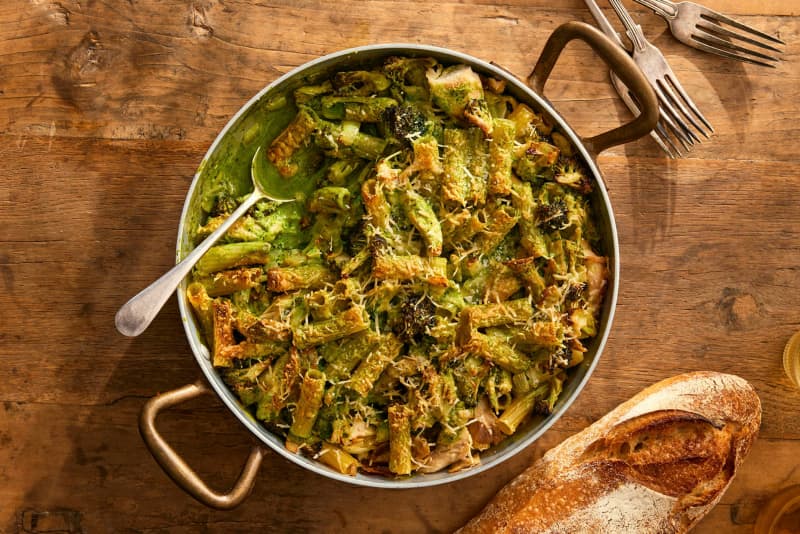 A round dish of chicken pasta bake on a wooden board, with forks and a baguette in shot.
