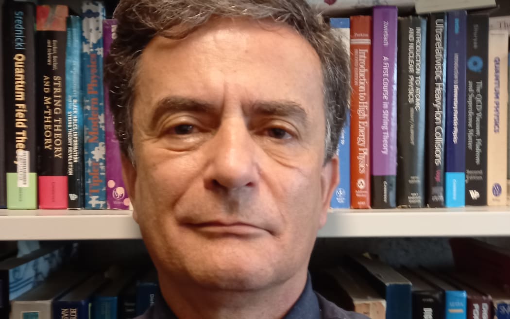 Dr David Krofcheck stands in front of a colourful bookshelf. He is wearing a dark collared shirt with a black tie. He is looking straight at the camera.