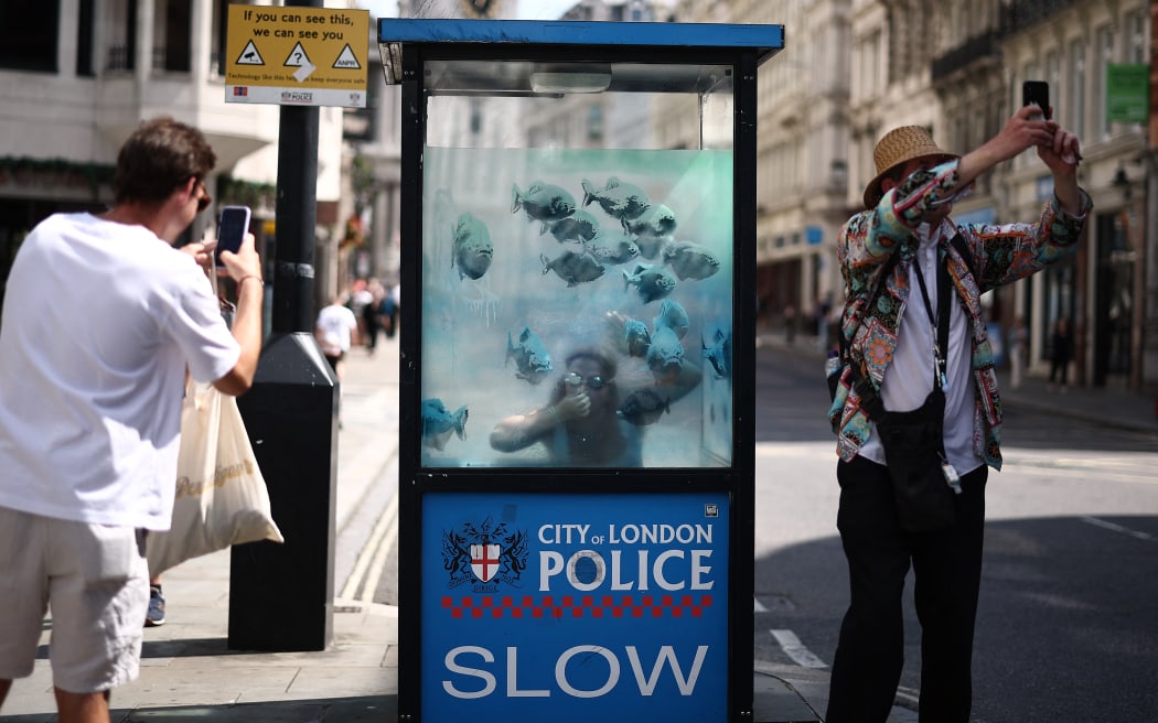 A person poses with an artwork by street artist Banksy, the seventh to released this week, depicting fish swimming around a Police Box, in the City of London, on August 11, 2024. The artist confirmed the work to be theirs after posting an image of it on the social media app Instagram. (Photo by HENRY NICHOLLS / AFP) / RESTRICTED TO EDITORIAL USE - MANDATORY MENTION OF THE ARTIST UPON PUBLICATION - TO ILLUSTRATE THE EVENT AS SPECIFIED IN THE CAPTION