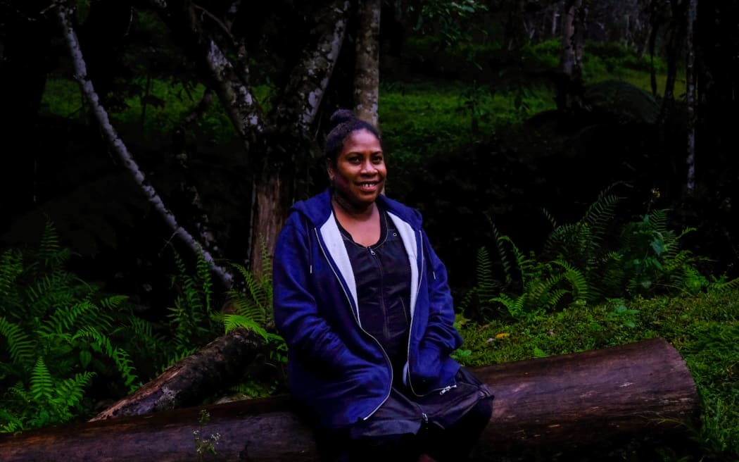 Yolarnie, a woman from Papua New Guinea, is wearing a black t-shirt and blue zip-up jacket. She sits on a log in front of a tree and ferns that feel dark and damp, like a rainforest. She is smiling and looking at the camera.