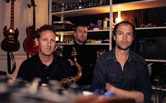 Love Square are a sat in what looks like an instrument storage room or maybe the control room of a small studio. There are guitars hanging on the wall behind them. One of them is holding a Saxophone.