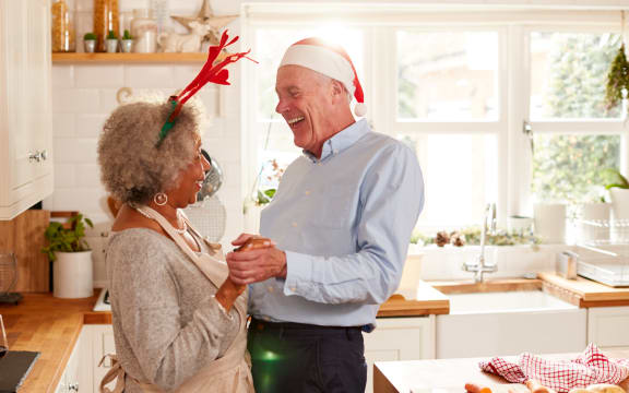 Loving Senior Couple Wearing Fancy Dress Antlers Dance In Kitchen Whilst Preparing Christmas Dinner