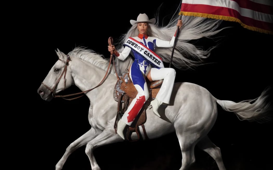 Cover image from Beyoncé's album 'Cowboy Carter'. Beyoncé sits side-saddle on a grey horse in red, white and blue outfit, cowboy hat and carrying the American flag.