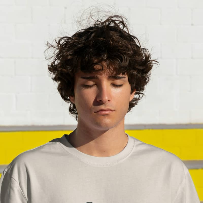 A boy with curly dark hair closes his eyes and stands against a white wall.