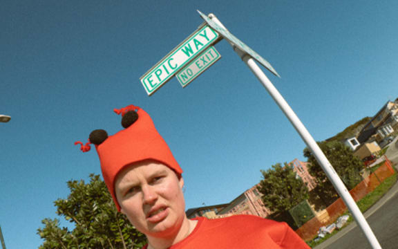 Comedian Liv Ward standing in a lobster costume in front of a street sign that reads "Epic Way", in Newlands, Wellington.