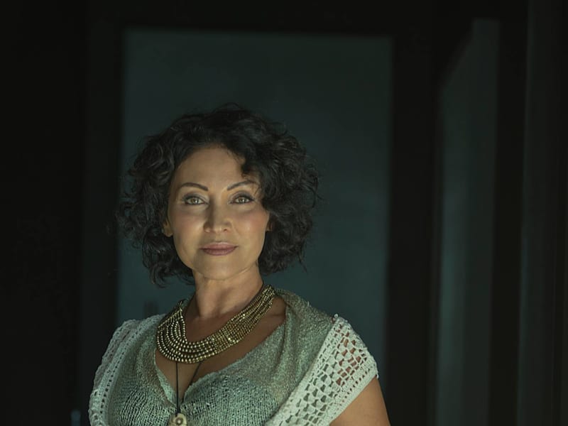 Tina Cross smiles slightly at the camera while clutching a knitted vest over a green dress.