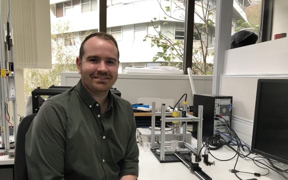 James sits next to a bench on which you can see a metal frame with an injector mounted vertically, a perspex box underneath into which skin is placed, and a track opposite this with a camera on it.