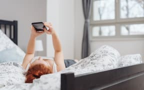 Young woman spending a relaxing day at home lying on her back on the bed reading a message on her mobile phone