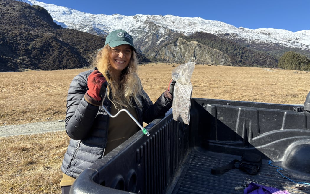 A woman with long blonde hair and a green cap holds up a plastic bag and a metal rod retrieved from the back tray of a ute. She is smiling and looking at the camera, wearing red gloves and a black puffer jacket. It's a blue cloudless sky above snow-capped mountains and a tussocked grassy valley floor.