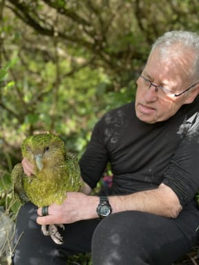 A man wearing a t-shirt and glasses sits in front of a tree, holding a large green parrot in his hands.