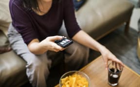 Woman sitting at home on couch, snacking, eating, and watching television.