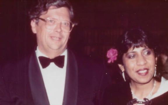 David and Jeya stand side by side, smiling. They are both in formalwear, David in a black suit jacket and black bowtie, and Jeya in a pink dress with a black cape over the top.