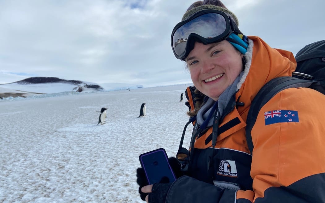 A woman wearing goggles pushed up onto her head smiles broadly at the camera. She is bundled up in a warm black-and-orange Antarctica New Zealand-branded jacket with a New Zealand flag patch on the arm. She has a backpack on and is holding a smartphone. Behind her on the ice, there are two small Adelie penguins waddling, and beneath a cloudy sky, there are snow-cloaked rolling rocky hills.