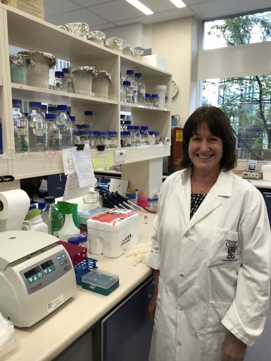 Tina Edgar, the Tate lab technician, stands by her lab bench which is full of glass bottles, plastic racks, desktop machines (centrifuge) and a Styrofoam box with samples in it.