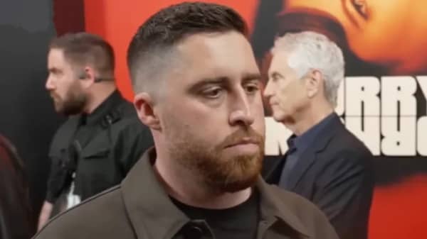 Trey Edward Shults is a bearded man wearing a brown jacket speaking to a reporter on a red carpet.