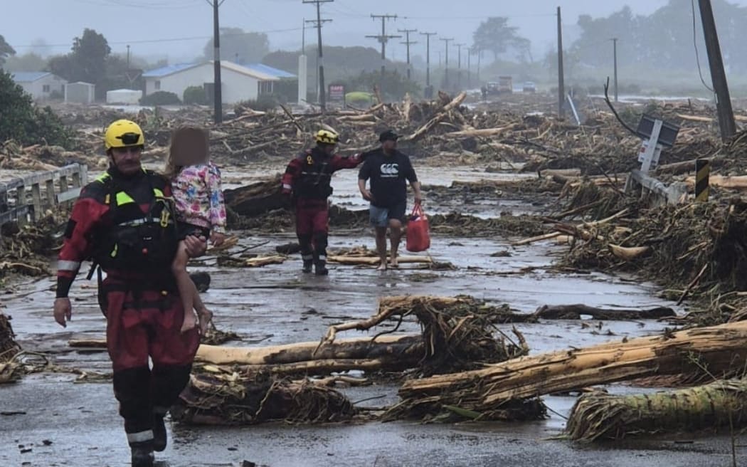 Firefighters, one carrying a young girl, assist residents down a flood hit road covered with woody debris