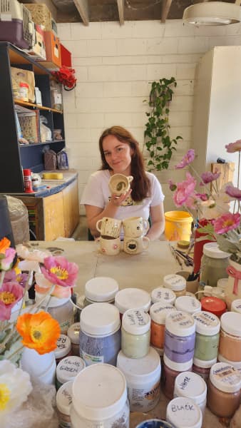Luni Pottery's Sam Peterson sitting at her pottery workshop with painted clay mugs and bunch of paint pots.