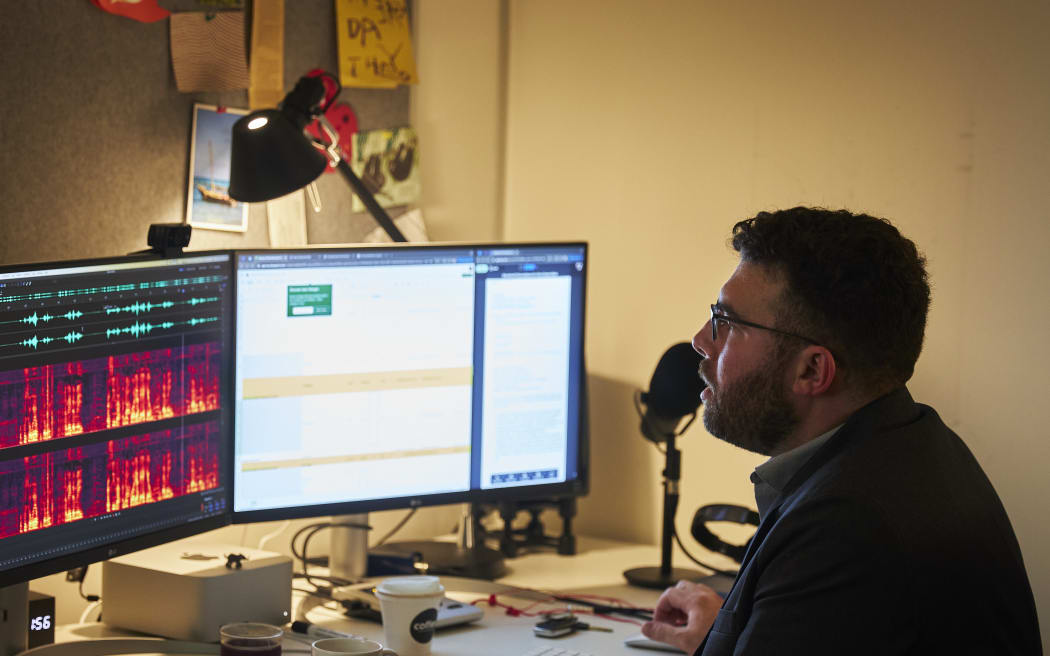 A man sitting at a desk looking at two computer monitors. One is showing brightly coloured waveforms. There is a microphone on the desk.