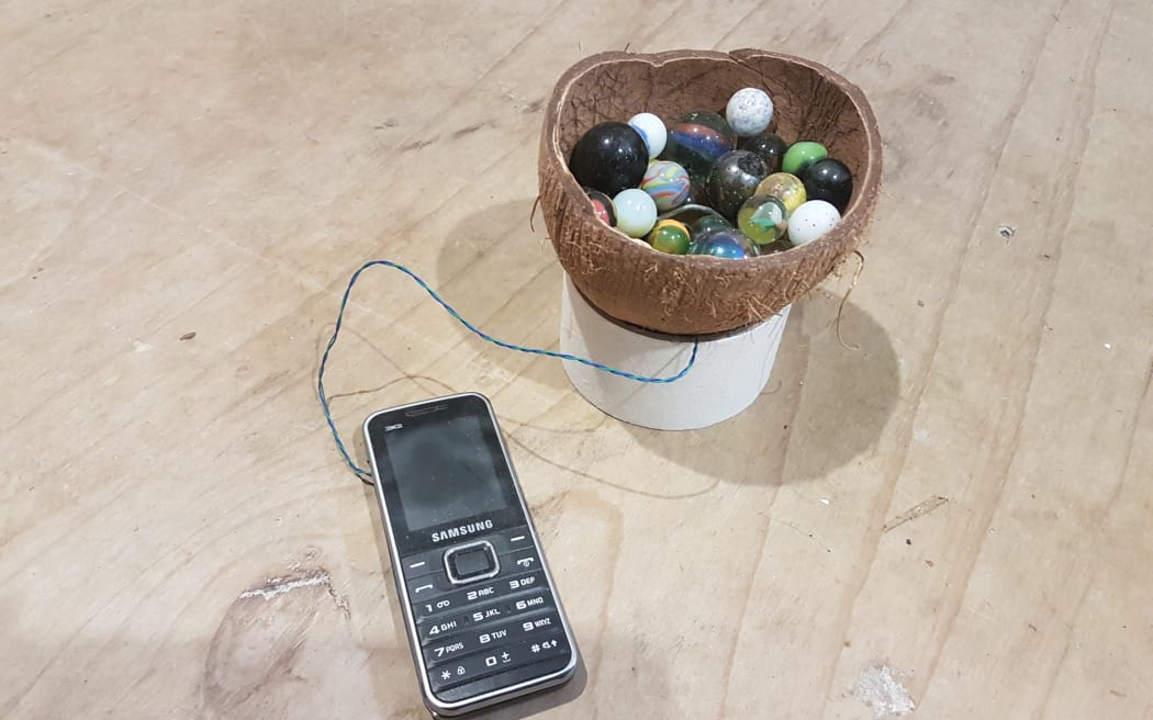 A old-school mobile phone is connected by a thin blue wire to a coconut shell, cut open, and filled with marbles