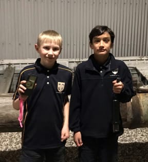 Young students Eddie and Austin hold up the hand held and hanging bat detectors. The hand held one is a small rectangle with a dial while the hanging bat detector is a microphone shape covered in black plastic.