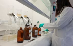 A woman wearing a white lab coat, rubber gloves and glasses reaches into a fume hood to grab a large brown glass bottle from a row of four similar bottles. Behind the bottles, there are three conical flasks with a filter funnel atop them, with brown-ish water filtering through.