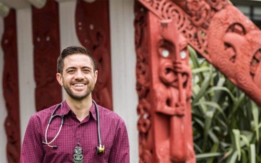 Dr Mataroria Lyndon, Senior Lecturer in Medical Education at the University of Auckland, stands in front of a wharenui wearing a button-up shirt smiling.