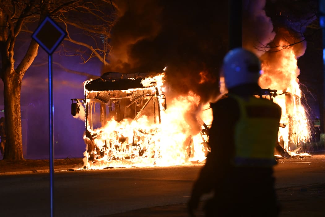 An anti-riot police officer standing next to a city bus burning during unrest in Malmo, Sweden.