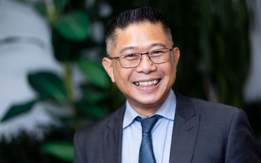 Mitchell Pham is wearing glasses with thin black frames, a suit with a blue shirt and tie. He is looking straight into the camera and smiling. His smile shows off his teeth. He looks genuinly happy. In the background is a large green plant in very soft focus.