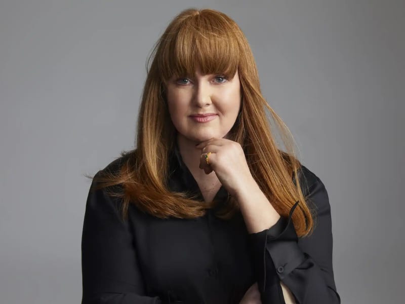 Megan Dalla-Camina - a white woman long red hair - smiles slightly at the camera and wears a black top.