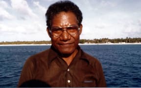 Portrait of Jeton Anjain wearing a brown shirt and wire rimmed glasses standing on a boat in front of the ocean and an island.