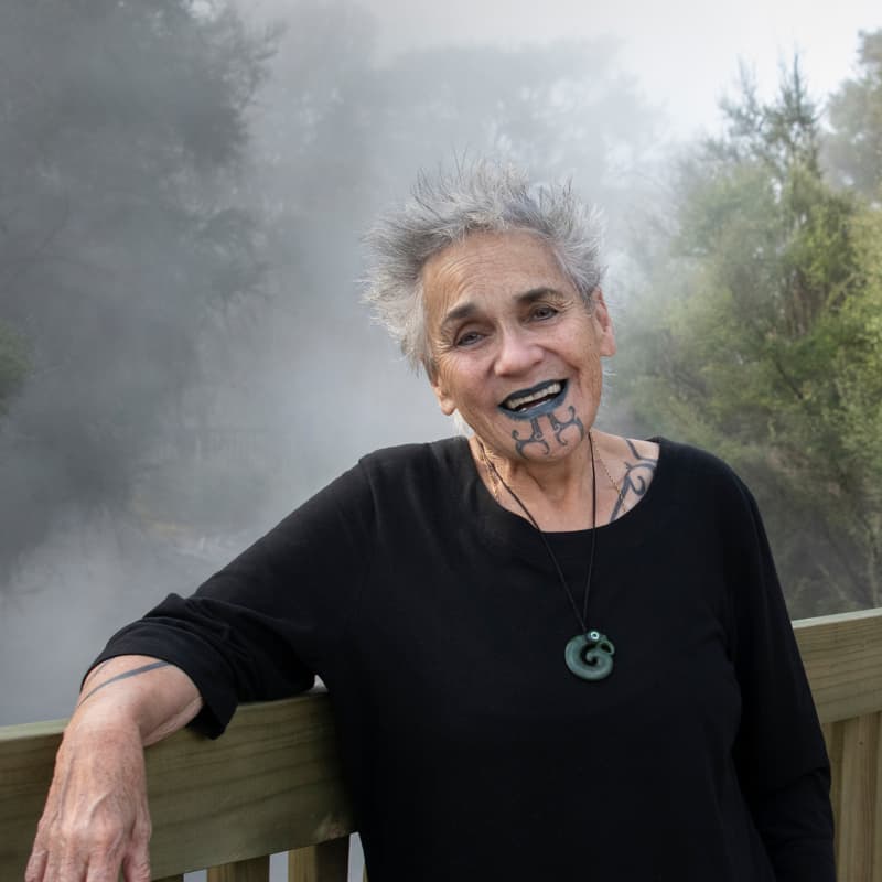 Ngāhuia te Awekōtuku (Te Arawa, Ngāpuhi, Waikato) stands by a wooden railing in nature.
