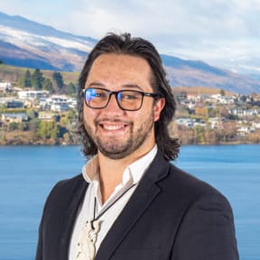 Marcus smiles at the camera. He is wearing a white shirt, black suit jacket, and a taonga around his neck. In the background is a view of a township on a blue lake, with mountains in the distance.