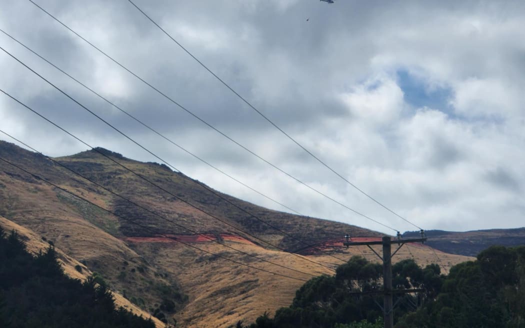 A helicopter holding a monsoon bucket flies over the Port Hills marked with red fire retardant at the back of Early Valley Road on 17 February, 2024.