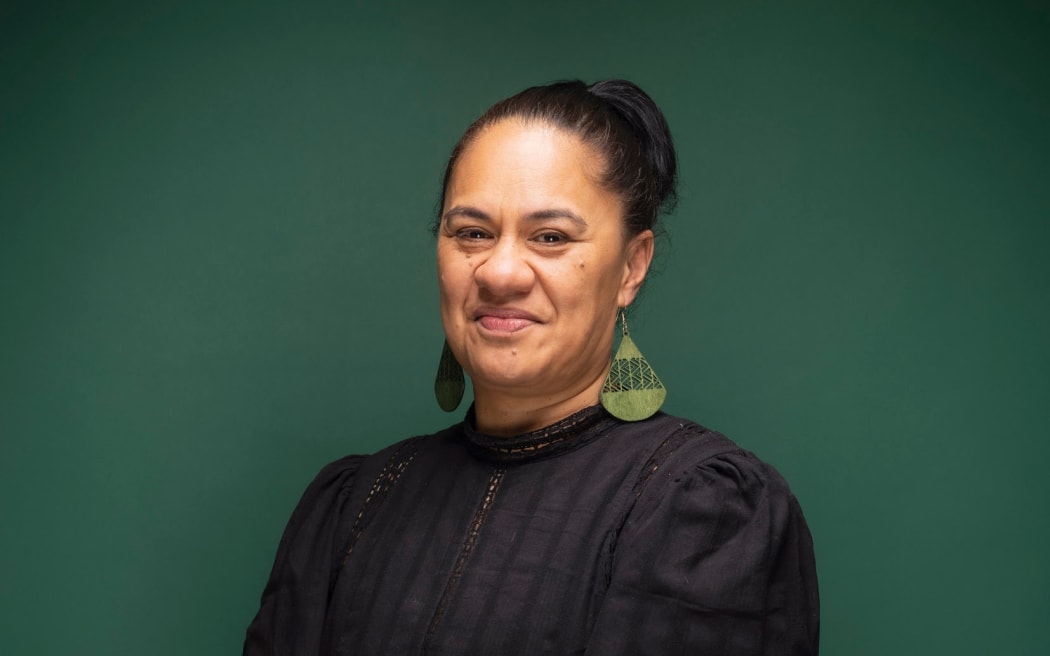 Hapai te Hauora Chief Executive, Jacqui Harema, stands smiling in front of a green backdrop wearing a black dress and green earings.