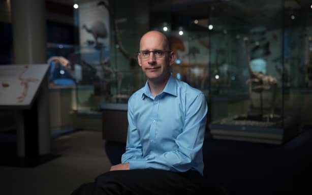 Portrait shot of Nic in a museum setting. He is wearing a blue long-sleeved shirt and glasses. He's sitting down looking towards the camera. Behind him are clear perspex display cabinets with Moa skeletons inside.