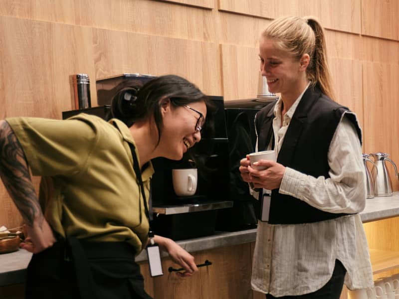 Two women smile next to a coffee machine.