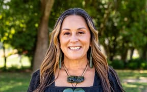 Kahira smiles at the camera. She is standing outside wearing a black top and a large taonga around her neck - a tiki, and a large pounamu heart.