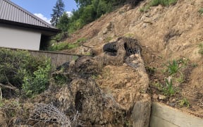 A hillside has collapsed into the back of someone's yard. A slip of loose earth wrapped in wire is slumped over a wooden fence. There is a neighbour's house and fence in the background. Above is a blue sky.