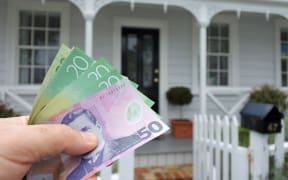 A mans hand holds NZ dollar bills against a front of a traditional villa house in Auckland, New Zealand. Buy, sale, real estate, insurance, mortgage, bank loans and housing market concept.