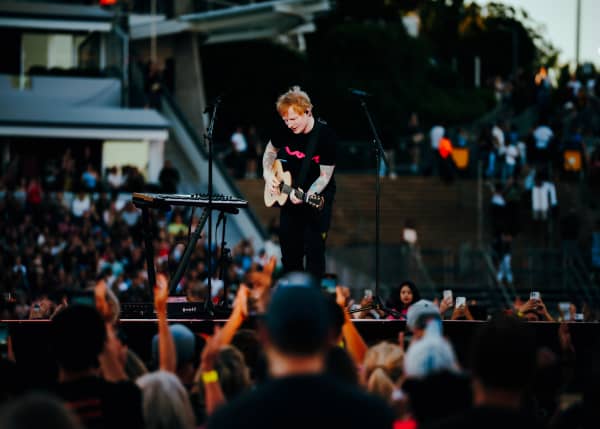 Ed Sheeran, wearing a black t-shirt, stands on stage playing a guitar.