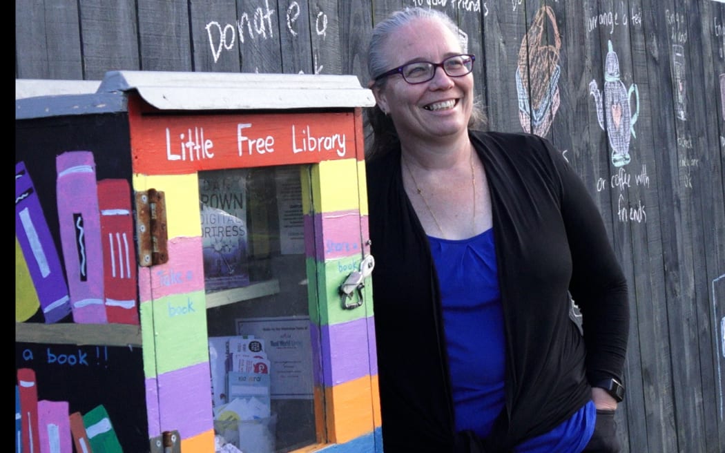 A woman smiles while standing next to a little free library.