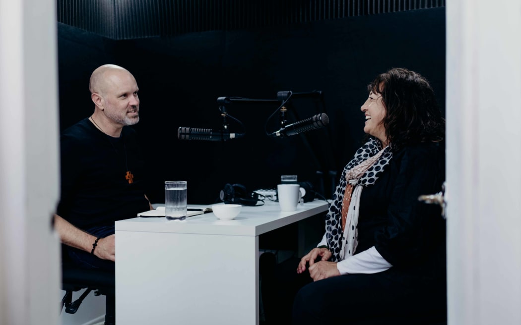 Reverend Frank Ritchie sits opposite Carmen Parahi in a small recording studio, mics in front of them as they discuss Carmen's career for the podcast re_covering.
