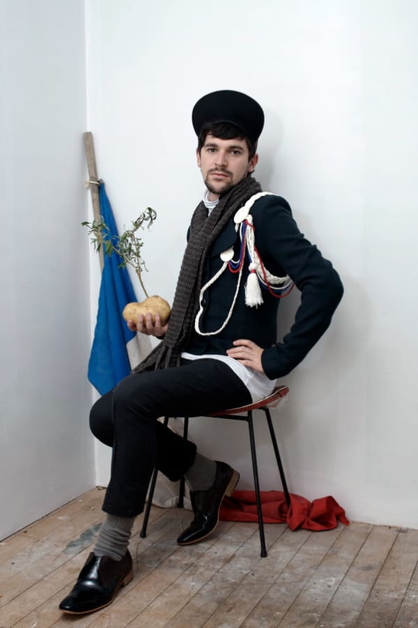 Dudley Benson wears a military jacket and hat and poses on a stool with one hand on his hip.