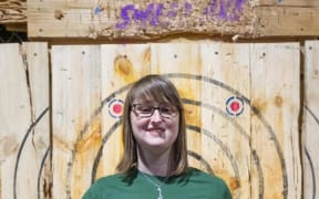 Britt stands in front of a target wall. She holds a trophy in one hand and an axe in the other.