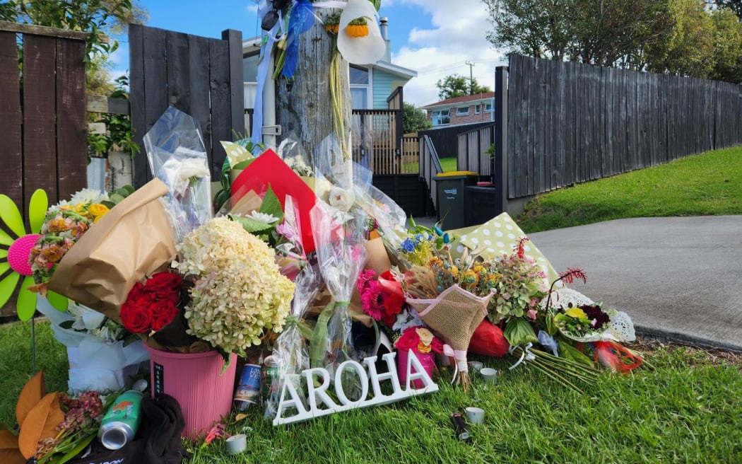 A pile of flower bouquets lay on the side of the road, on grass, and a letters spell out aroha.