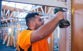 A man in a fluorescent orange shirt drills a nail into a structure. He is standing in a construction site.